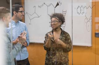 Three university students work on a chemistry assignment in the lab. One is writing with a marker on a transparent glass board.